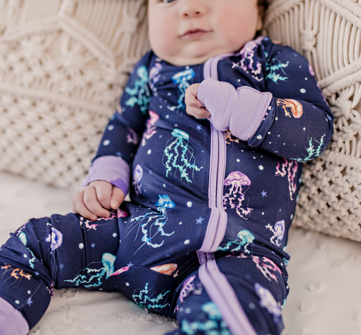 Baby wearing one piece pajama with dark blue background and purple cuffs with maine jellyfish in blues, teals, purples, and pinks