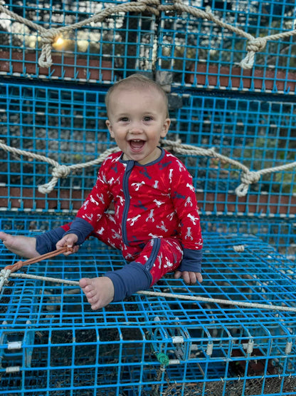 A baby wearing a red and blue maine lobsters Abel Zippy Romper, sitting on a blue lobster trap