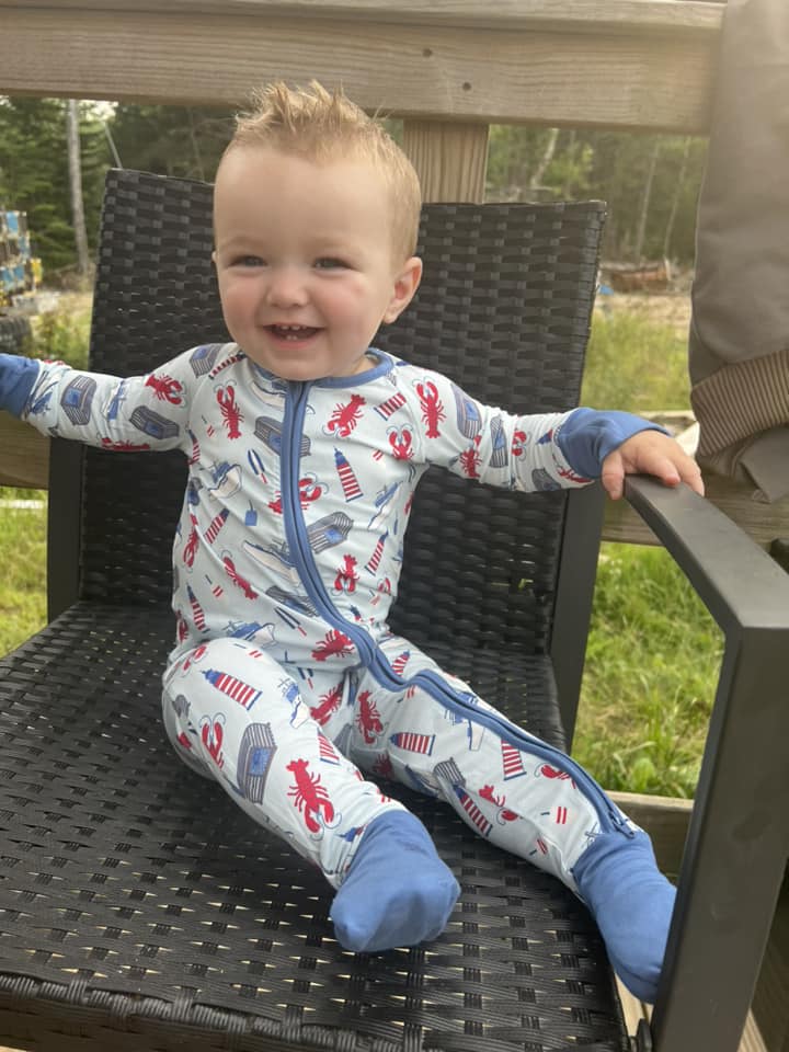 Child wearing a one piece zippy romper with a light blue background and darker blue cuffs with red lobsters, white and blue boats, blue, white, and red lighthouse, gray and blue traps, and red, white, and blue buoys set in the Atlantic Ocean off the coast of Maine