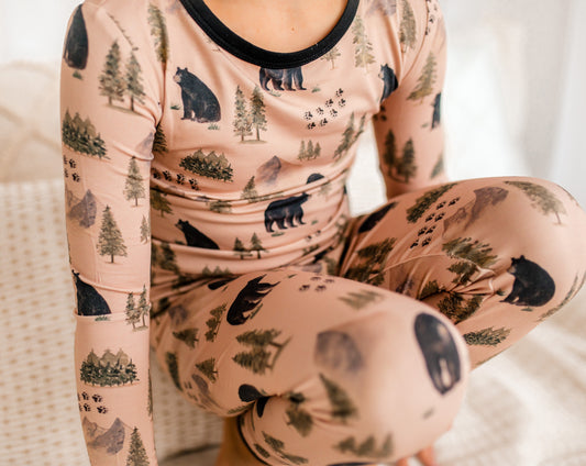 Child wearing a light brown pajama set with Maine black bear and bear tracks, along with green trees and gray mountains. 