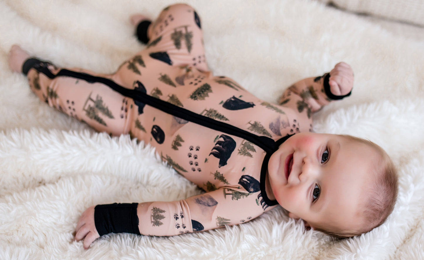 Baby wearing a tan one piece with black bears and bear tracks along with green trees from the Maine forest 