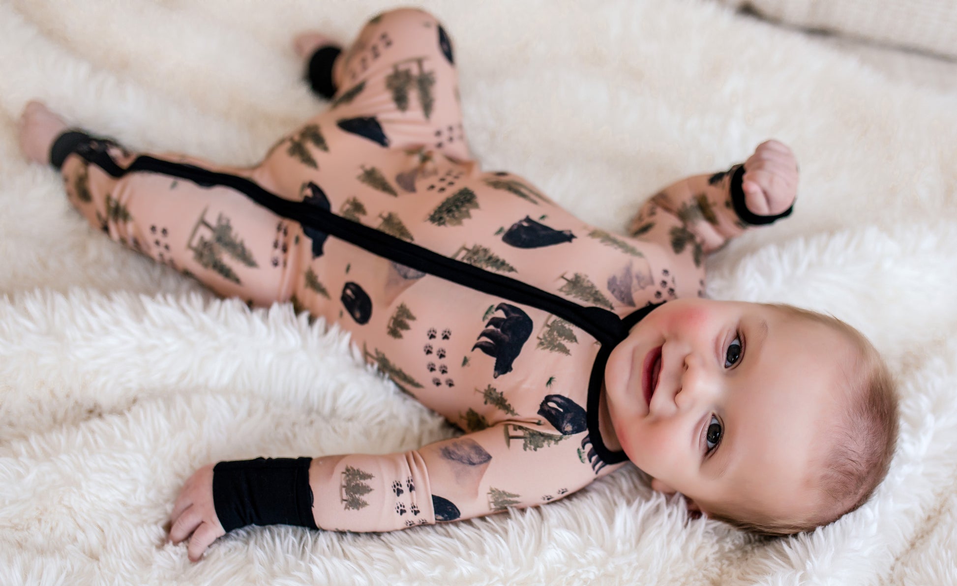 Baby wearing a tan one piece with black bears and bear tracks along with green trees from the Maine forest 