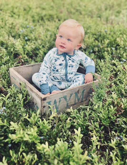 A baby sitting in a wooden crate wearing a blue and white romper with blueberries from Maine all over 
