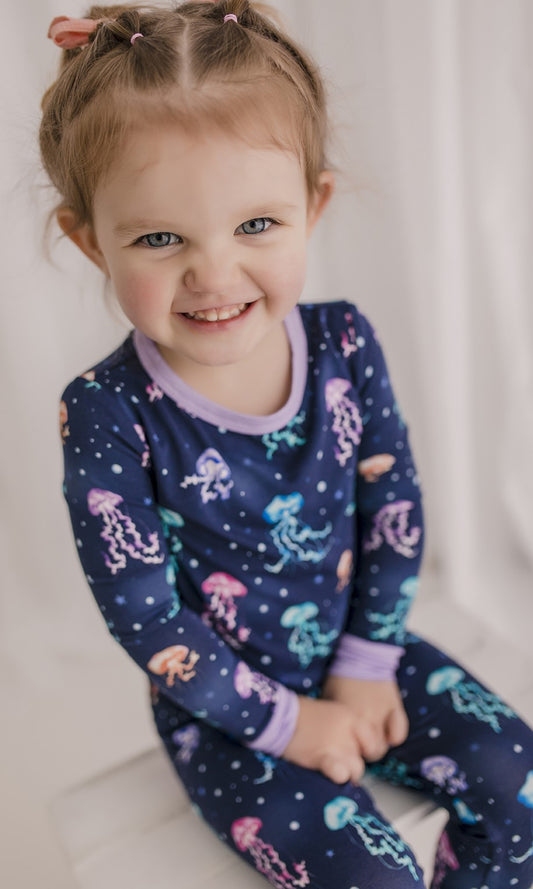 A toddler wearing a two-piece pajama set with a pattern of jellyfish in shades of blue, purple, teal, and orange with purple cuffs 