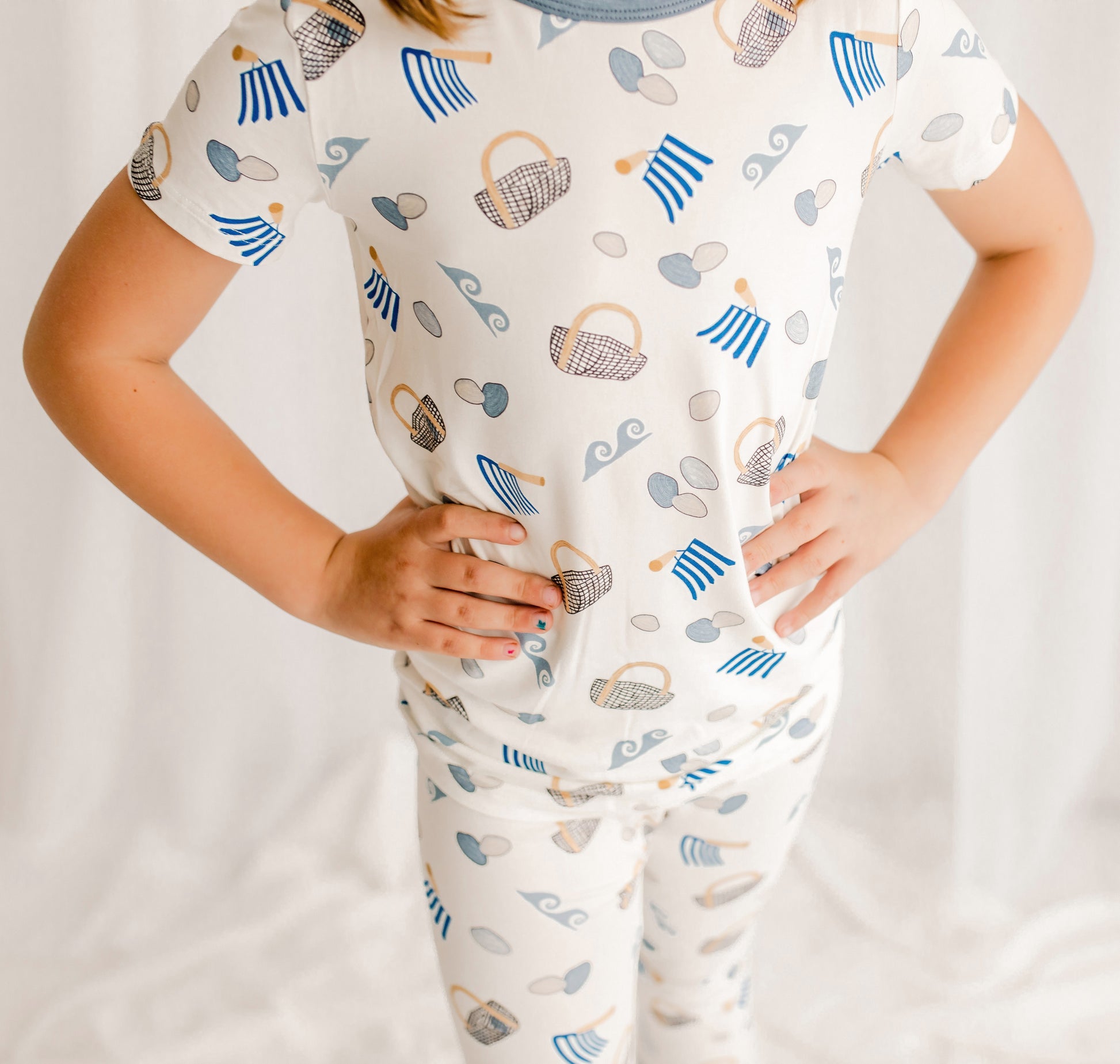 Child wearing two piece pajama set with clams, baskets, diggers, and waves with blue, gray, and tan accents on a white background. 
