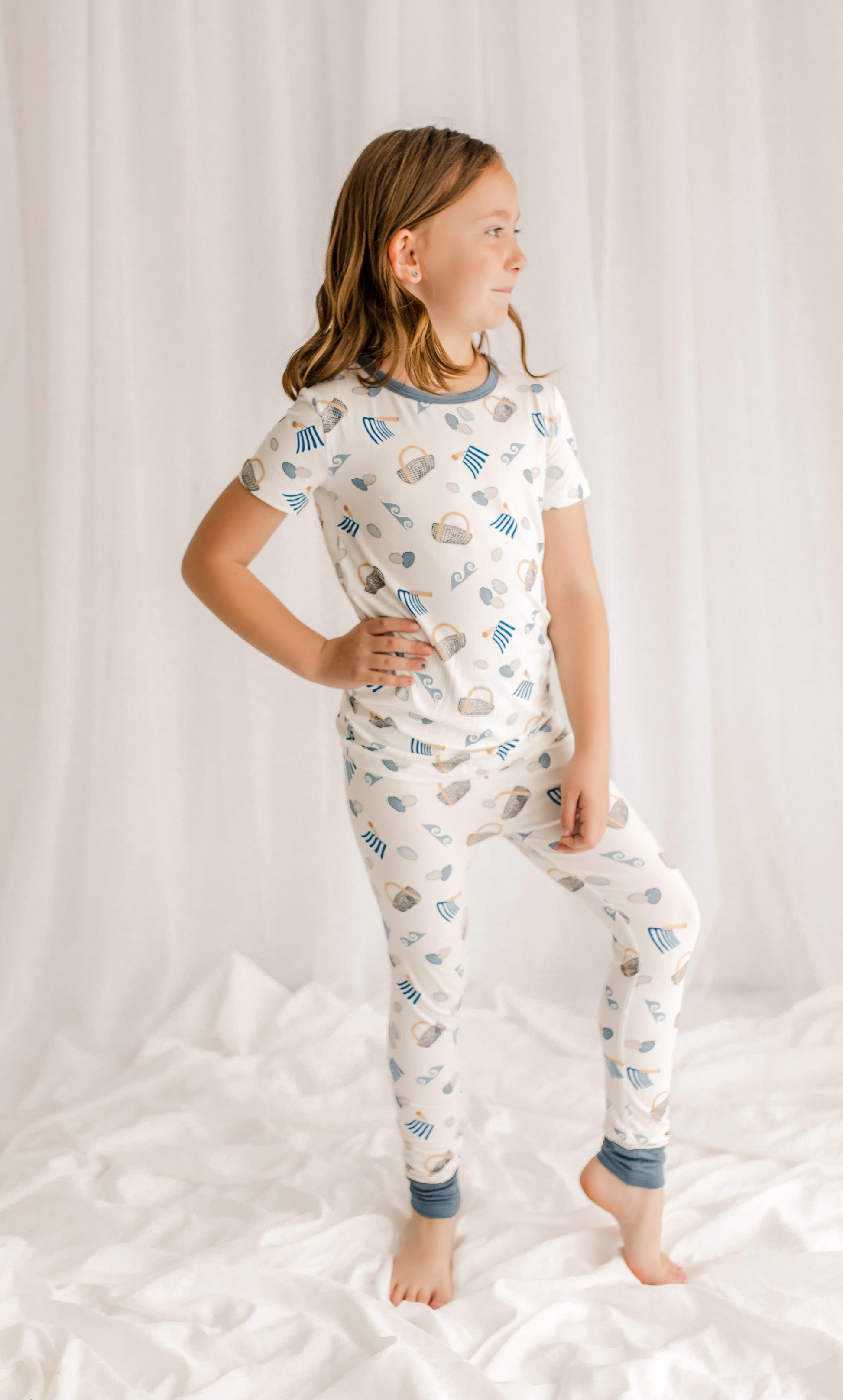 Child wearing two piece pajama set with clams, baskets, diggers, and waves with blue, gray, and tan accents on a white background. 