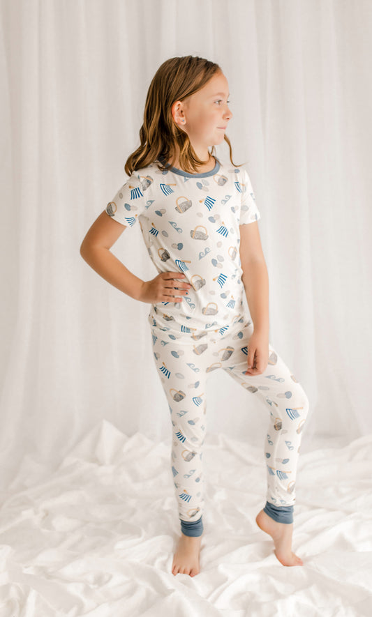 Child wearing two piece pajama set with clams, baskets, diggers, and waves with blue, gray, and tan accents on a white background. 