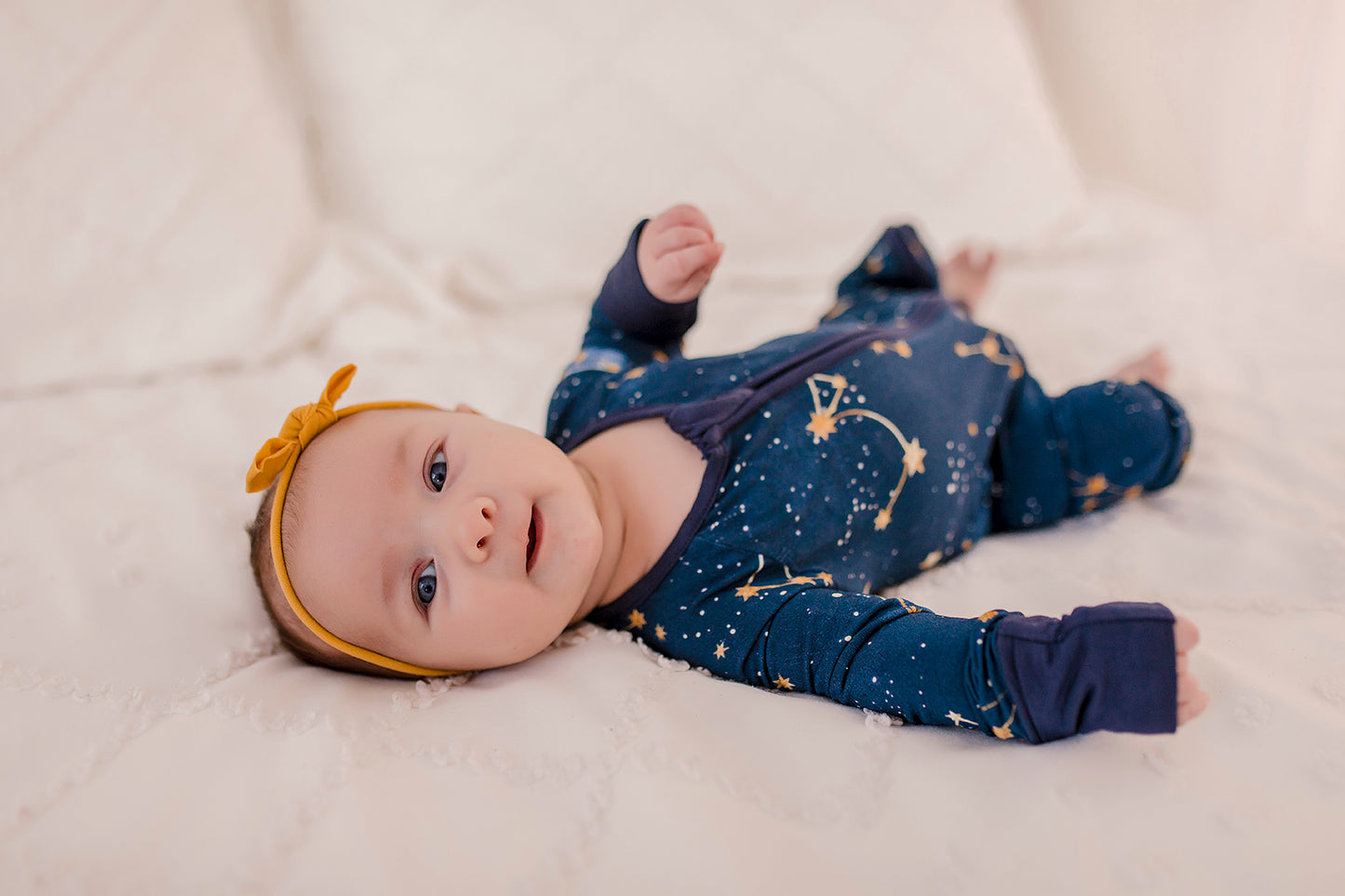 A baby wearing a dark blue romper with gold dots and stars that make up constellations in the maine sky with blue cuffs 