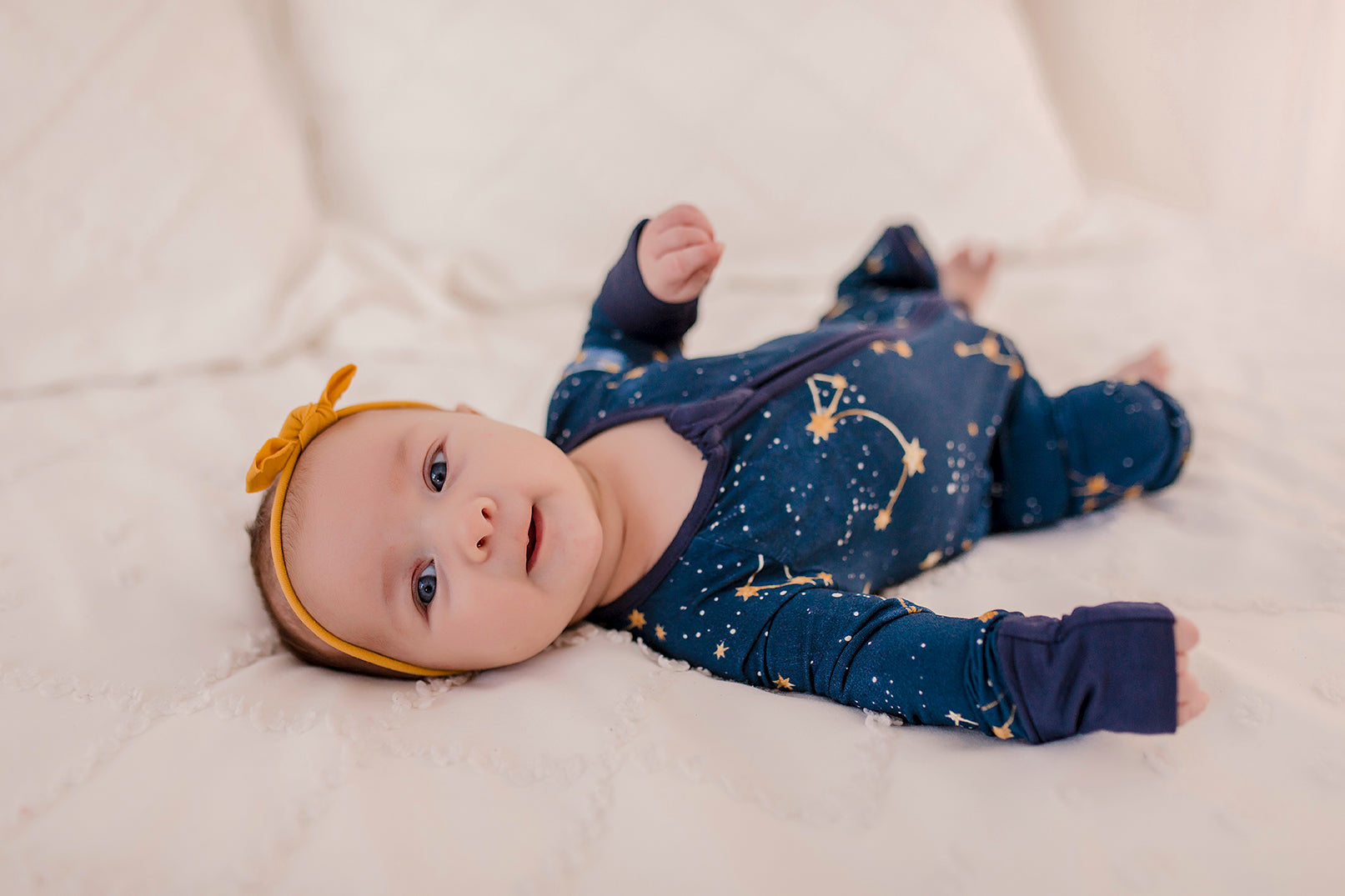 A baby wearing a dark blue romper with gold dots and stars that make up constellations in the maine sky with blue cuffs 