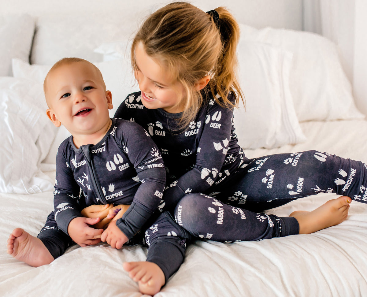 Two children in matching gray pajamas with white lettering and woodland animal tracks sitting on a bed.