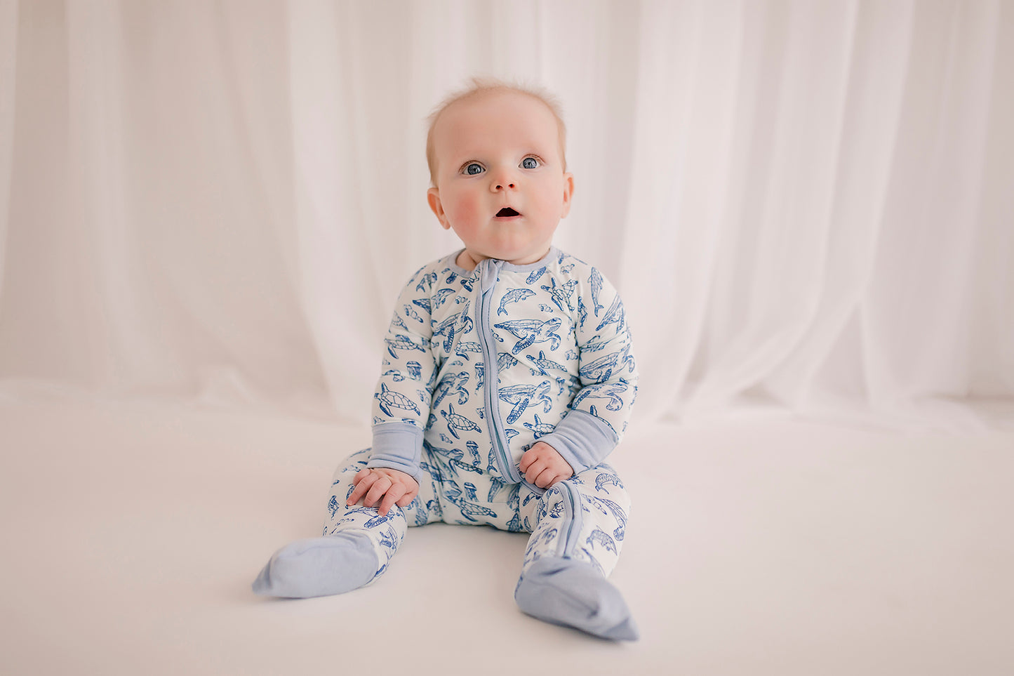A baby wearing a long-sleeved romper with an ocean animal pattern that has sea turtles, dolphins, and jellyfish, in shades of blue