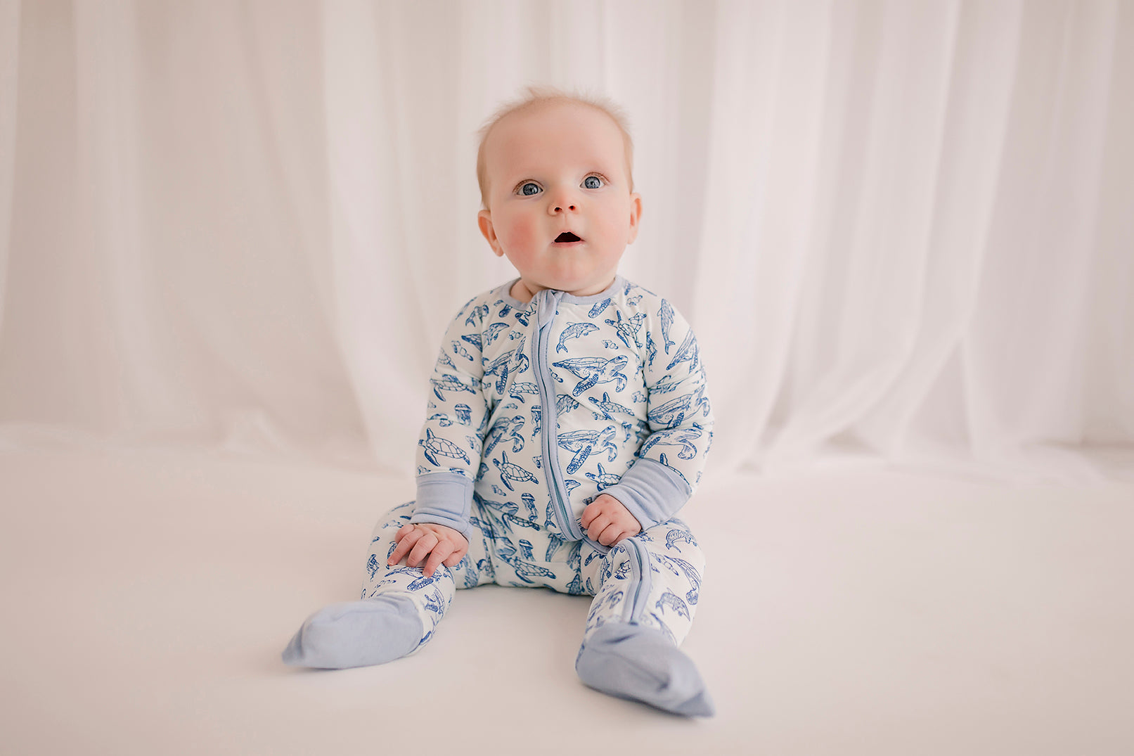 A baby wearing a long-sleeved romper with an ocean animal pattern that has sea turtles, dolphins, and jellyfish, in shades of blue