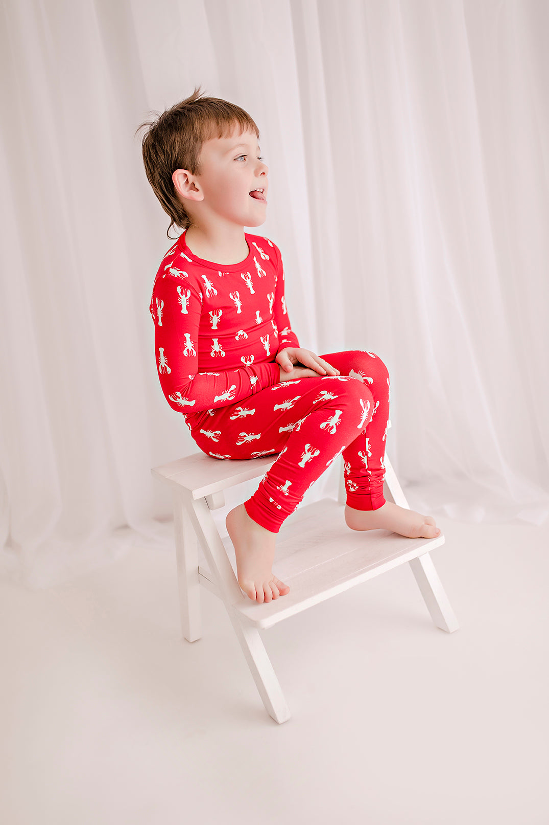 Child wearing coastal maine themed pajamas with long sleeves and pants on a red background with white lobster pattern 
