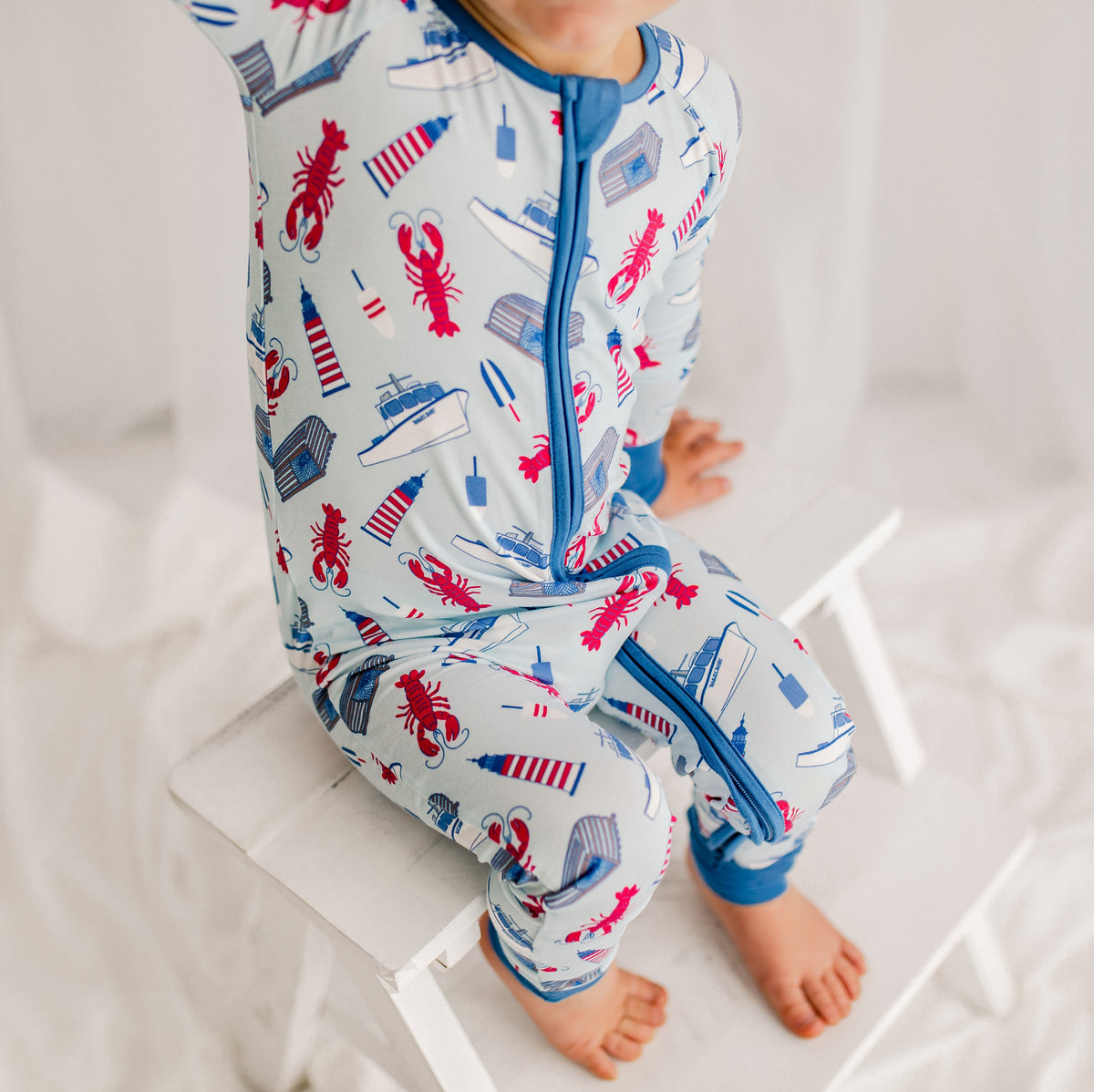 Child wearing a one piece zippy romper with a light blue background and darker blue cuffs with red lobsters, white and blue boats, blue, white, and red lighthouse, gray and blue traps, and red, white, and blue buoys set in the Atlantic Ocean off the coast of Maine