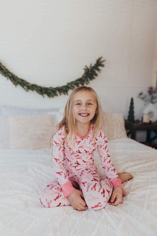 A child sitting on a bed wearing a two-piece pajama set with a pink  Maine Atlantic Ocean lobster design.