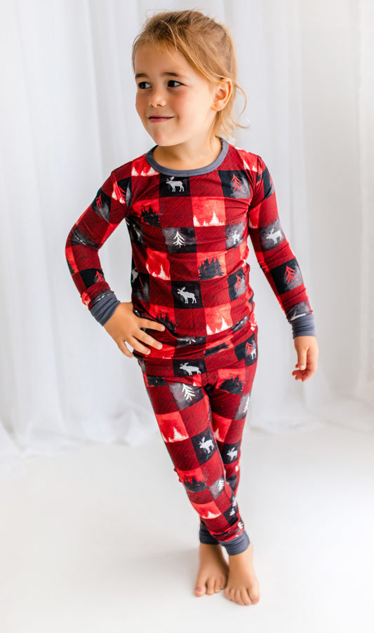 Child wearing long sleeved shirt and pants with red and black plaid squares with different kinds of trees and a moose inside the squares 