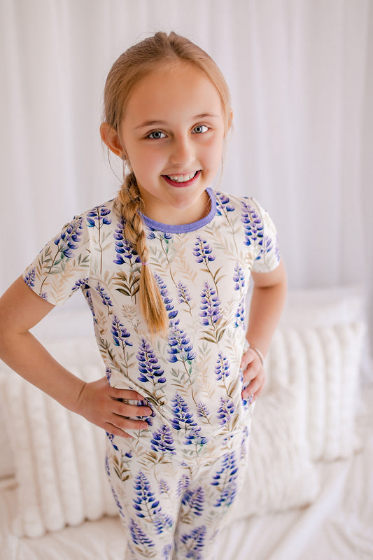Girl wearing a floral pajama set against a white background that has purple lupines all over 