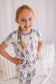 Girl wearing a floral pajama set against a white background that has purple lupines all over 