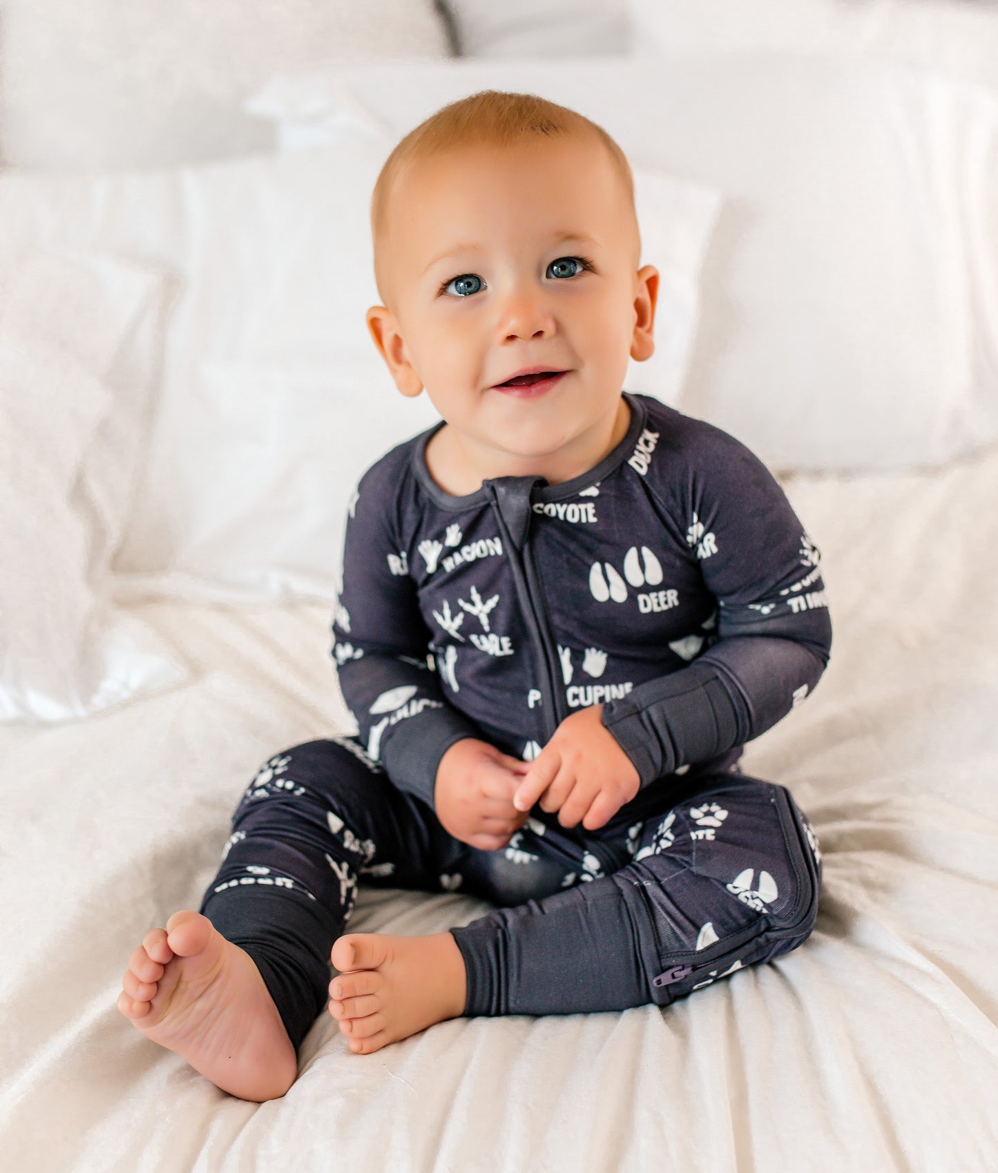 Baby wearing a dark gray bamboo onesie with white patterns and lettering sitting on a white blanket.
