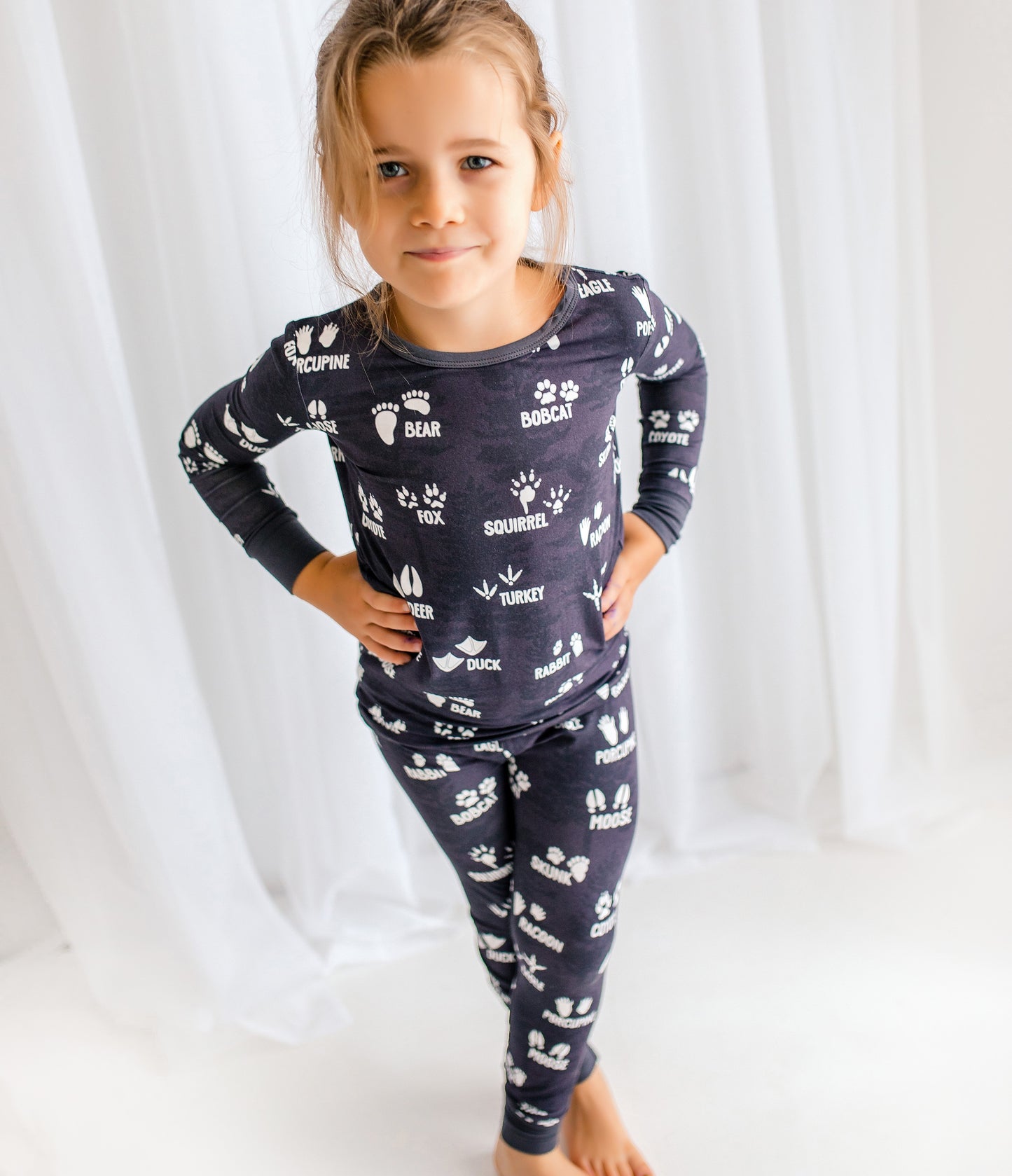 Child standing wearing dark gray two piece pajamas with white animal prints and lettering with a white background