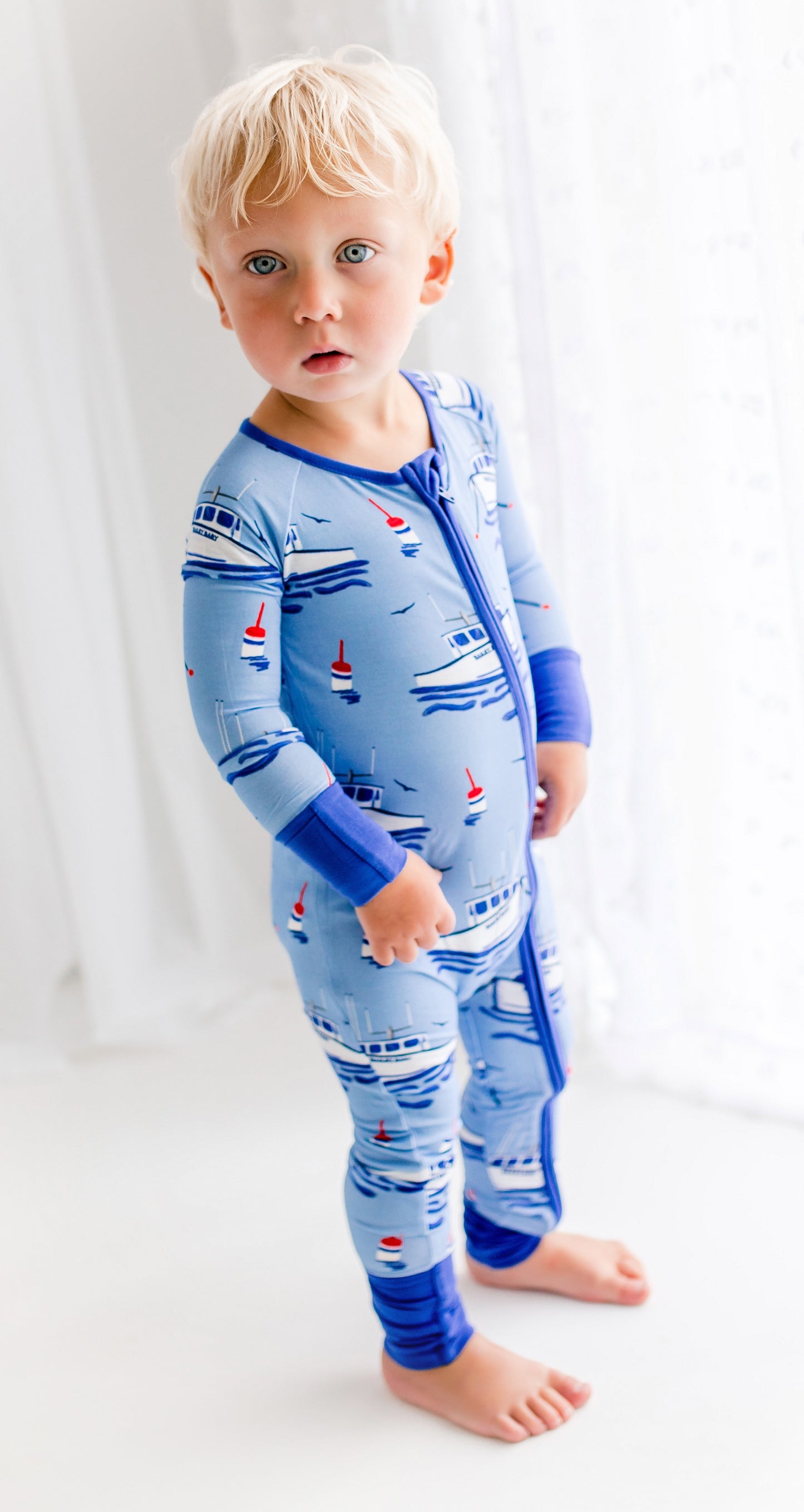 Child wearing one piece pajama set with boat patterns and buoys on a light blue background 