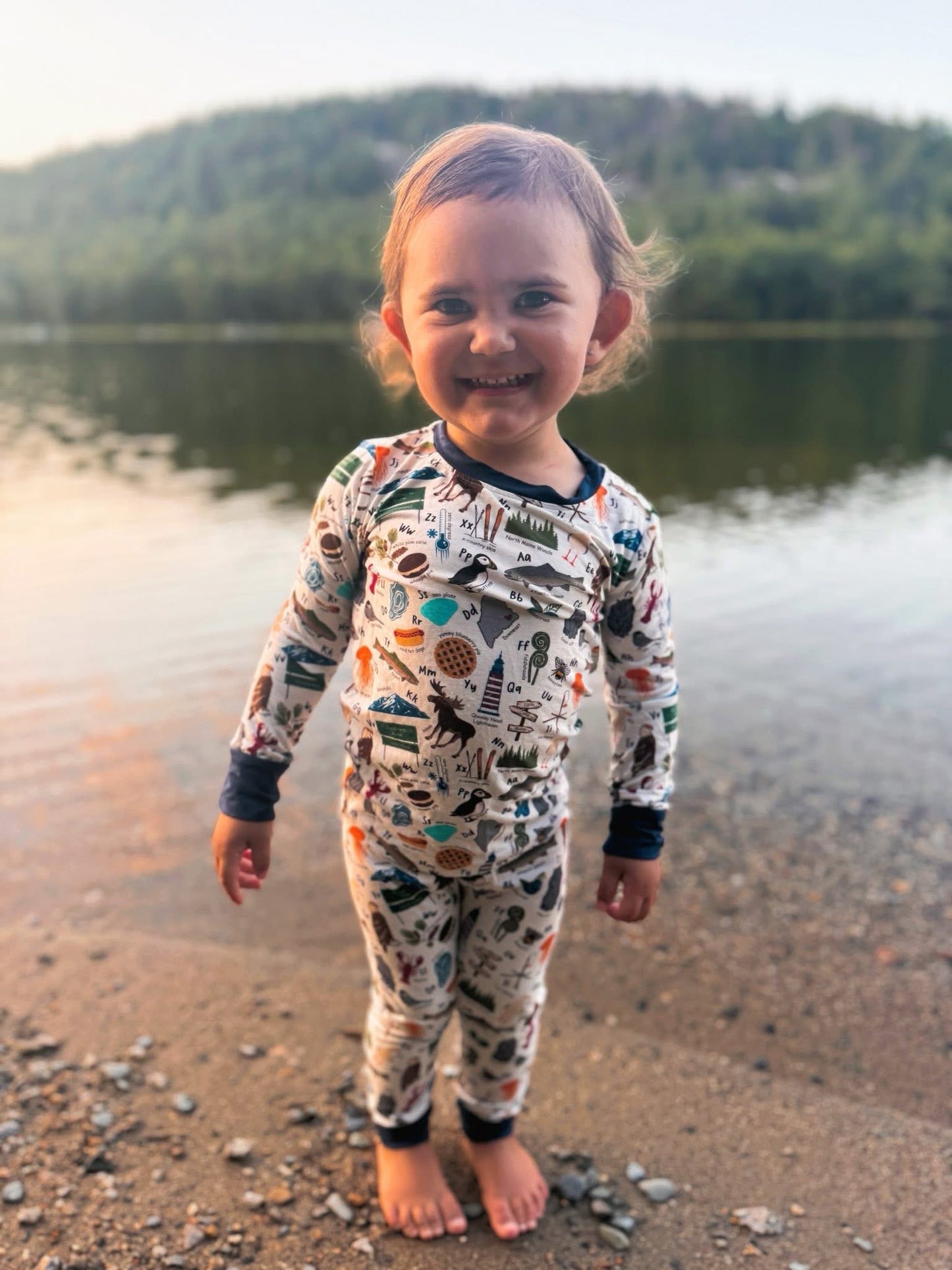 Child wearing an alphabet themed one piece pajama about all the things that represent maine, like puffins, moose, blueberries, eagles, and lobsters on a white background with blue cuffs. 