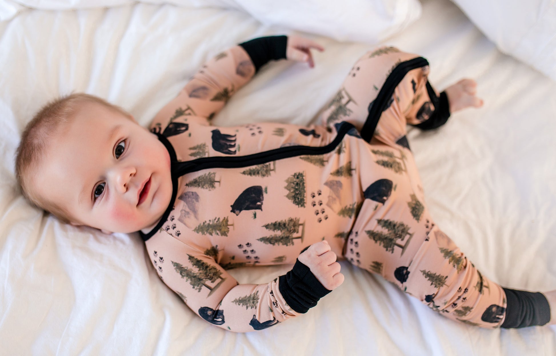 Baby wearing a onesie with Maine black bears and trees, mountains and bear tracks on a tan background 