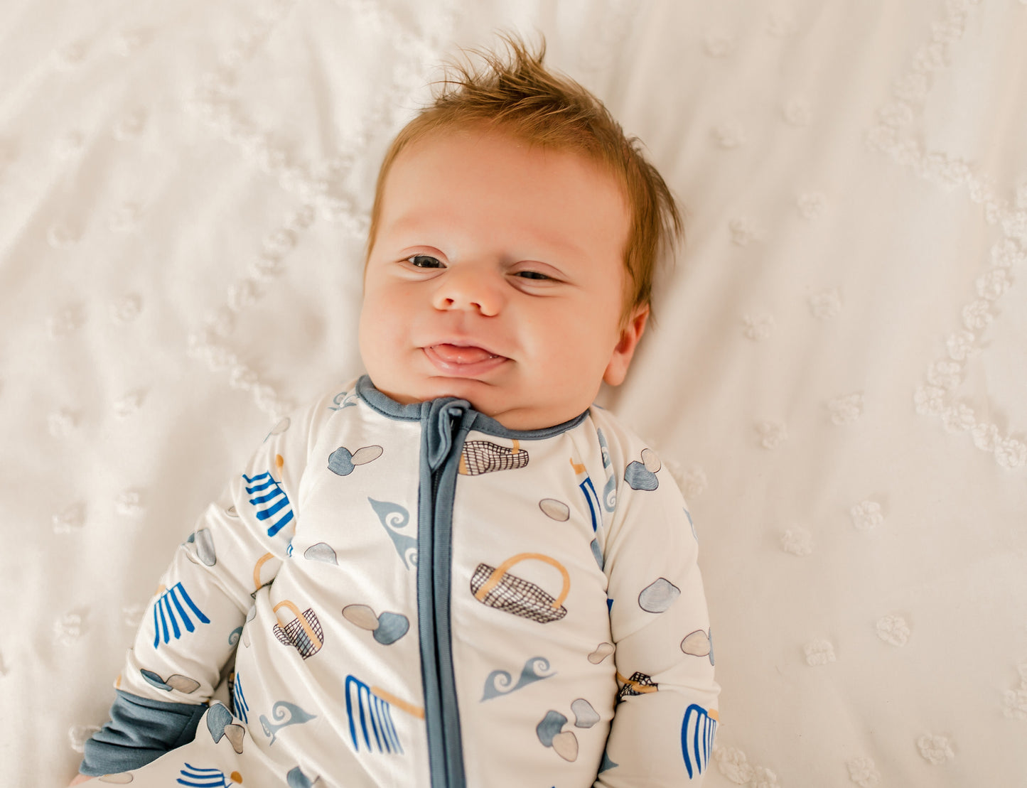 Baby wearing a zippy romper with blue gray and tan colors along with clams, diggers, baskets, and waves 