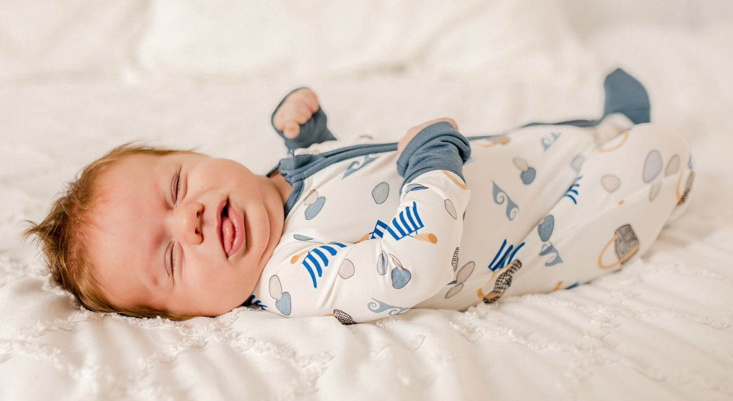 Newborn baby wearing a onesie with blue, gray, and tan prints of clams, baskets, waves, and clams on a white background. 