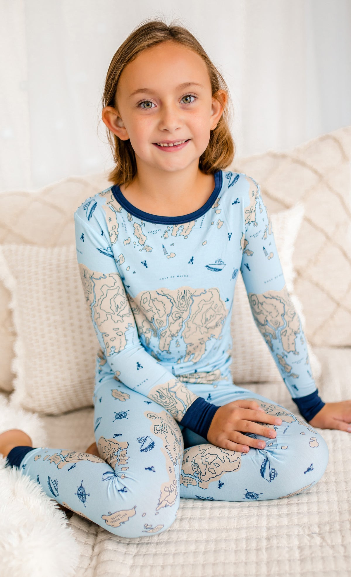 Child wearing a blue pajama set with a cream colored nautical map of a piece of the beautiful Maine coast, blue lobsters, blue lobster boats, and blue compasses. 