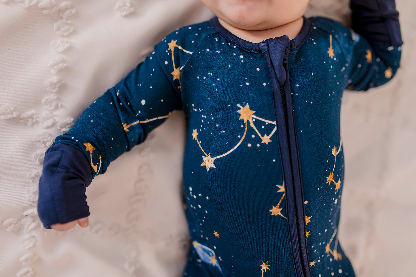 A baby wearing a dark blue romper with gold dots and stars that make up constellations in the maine sky with blue cuffs 
