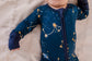 A baby wearing a dark blue romper with gold dots and stars that make up constellations in the maine sky with blue cuffs 