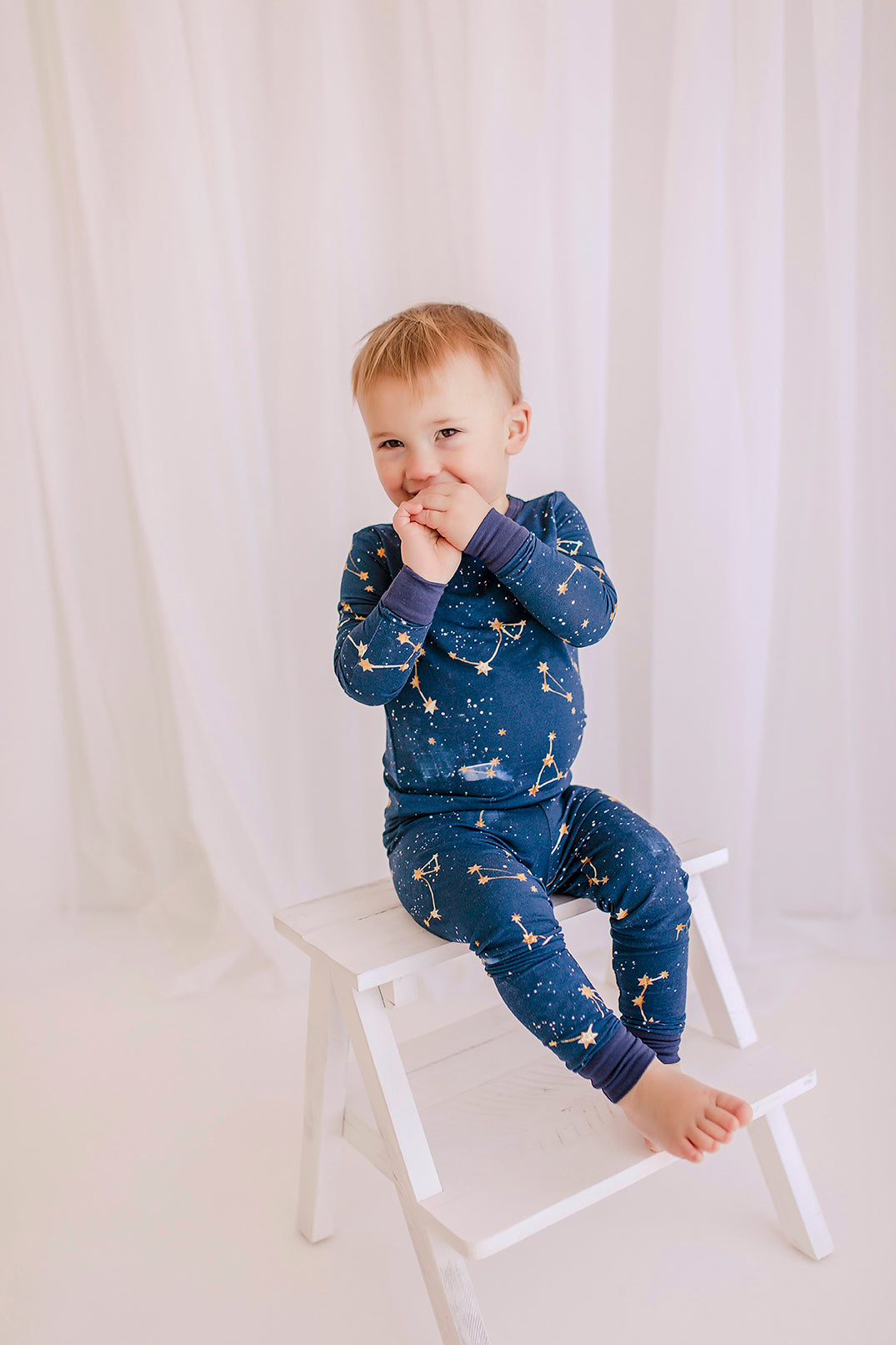 Child wearing long sleeved two piece pajama set on a dark blue background with constellations, galaxy, and stars in yellowish gold with blue cuffs 