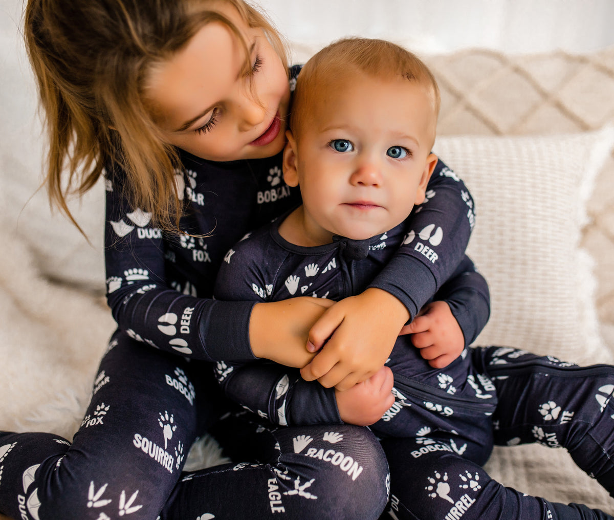 Two children in matching pajamas sitting on a couch with forest animal tracks in white and a dark gray background 