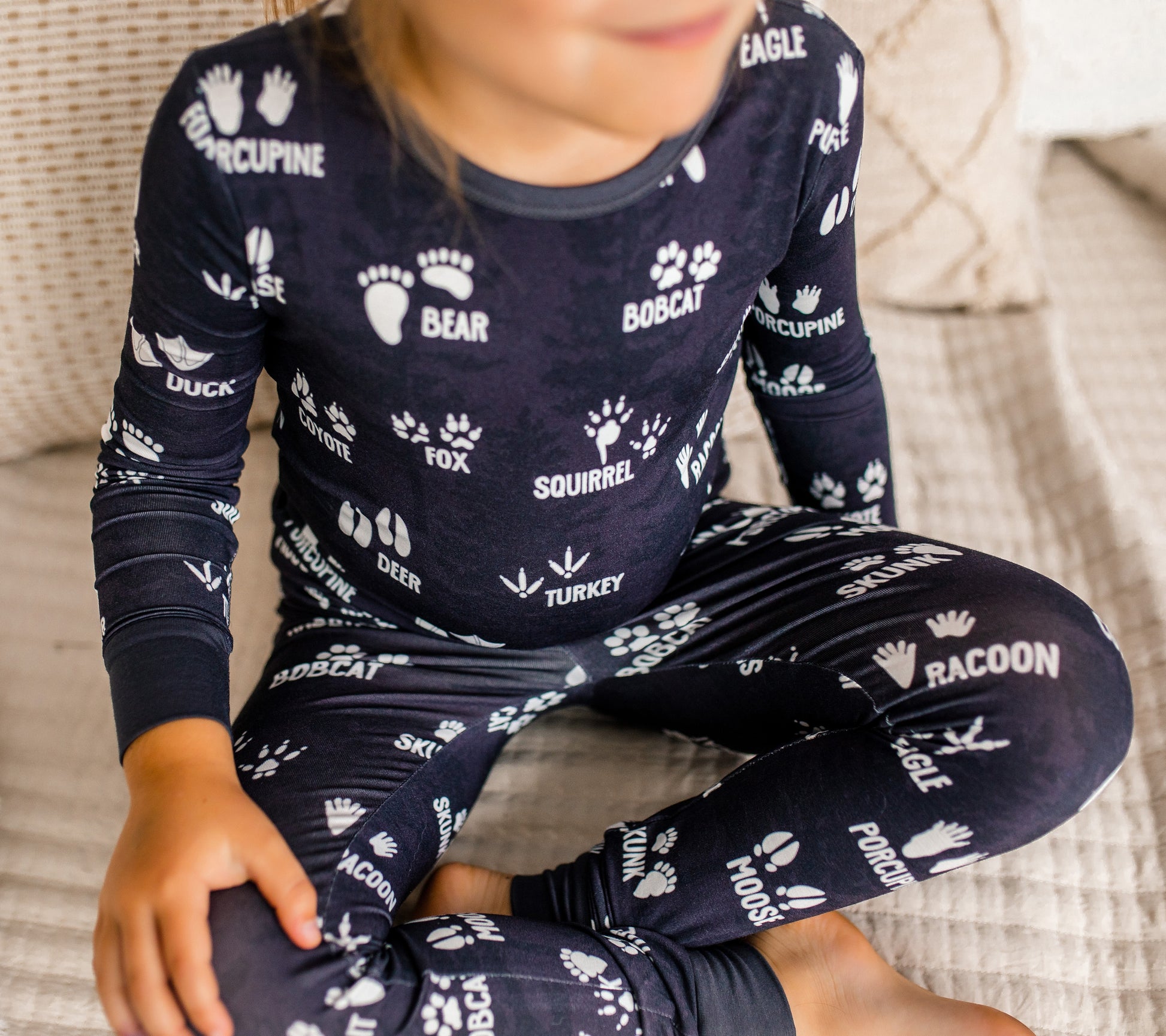 Child wearing gray two piece organic bamboo pajamas with white animal prints sitting cross legged on a couch