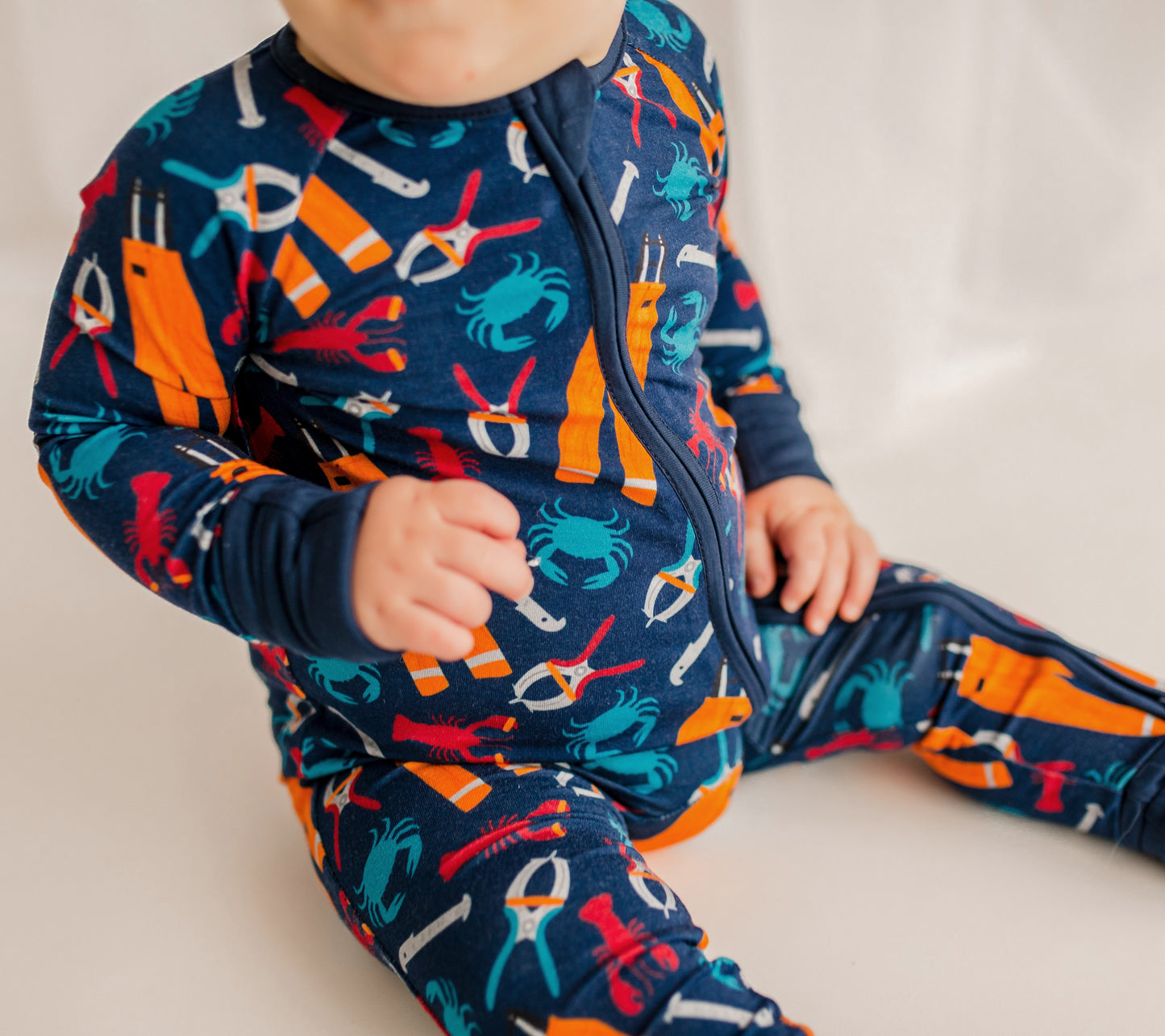Child wearing a navy blue outfit with red lobsters and blue crabs and all the essentials to work on a fishing boat,  measure, bander, and oil gear. 