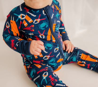 Child wearing a navy blue outfit with red lobsters and blue crabs and all the essentials to work on a fishing boat,  measure, bander, and oil gear. 