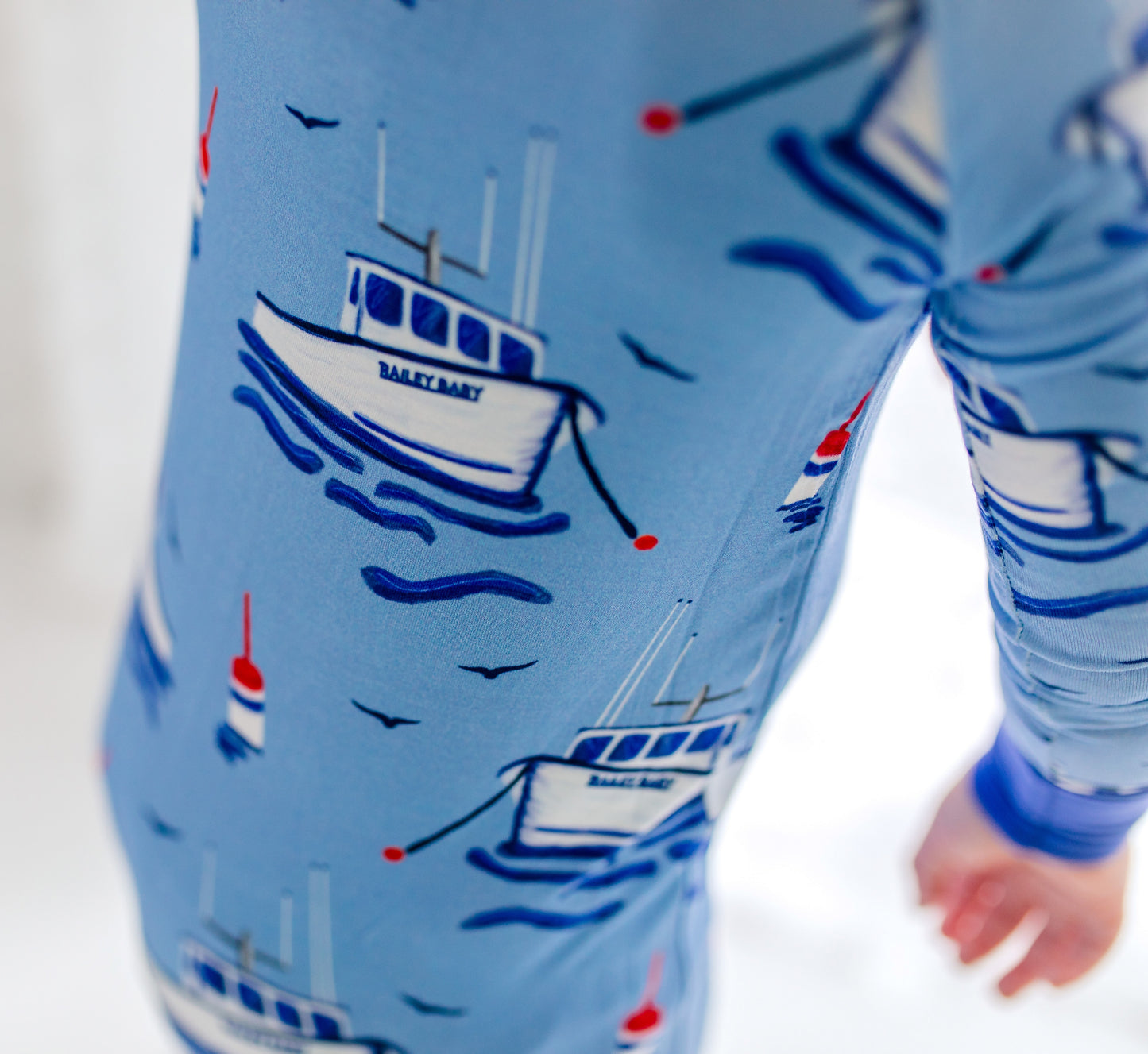 Children's light blue onesie with darker blue accents with fishing boats, floating red blue and white buoys, flying seagulls, and waves under the boats. 