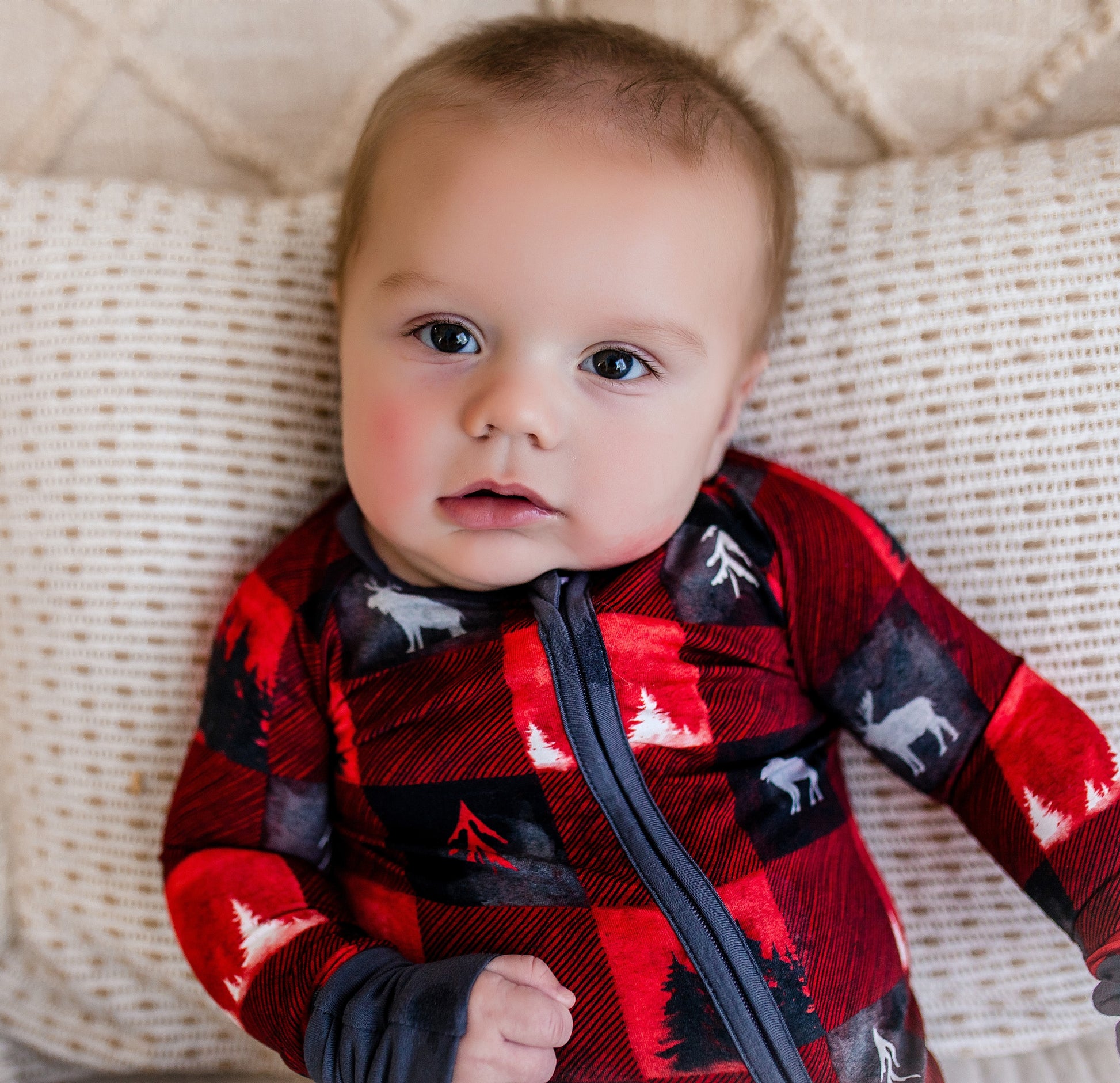 Baby wearing a red plaid zippy with images of black trees, white trees, and the largest animal in Maine, the moose. 