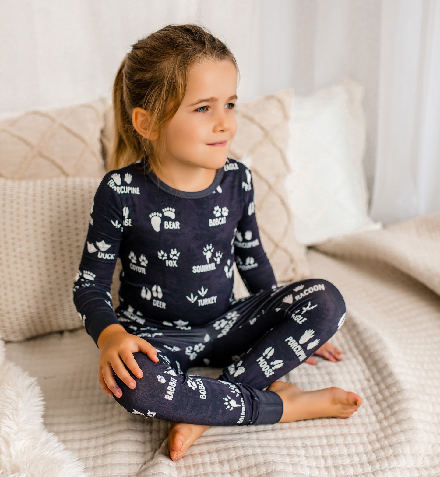 Child wearing gray pajamas with white text and tracks from all different wild animals, sitting on a bed