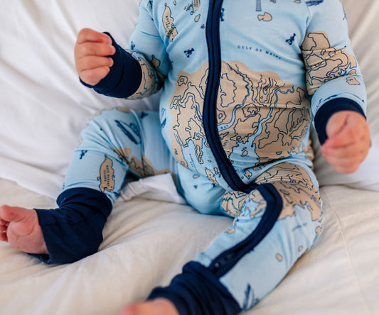 Baby in a blue and beige nautical map-themed one piece zippy of the vast maine coast with dark blue accents and nautical symbols such as lobster boats, lobsters, and compasses. 