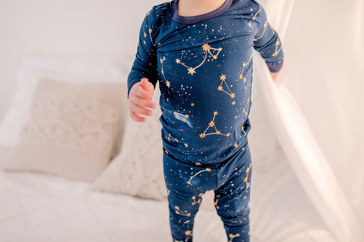 Child wearing long sleeved two piece pajama set on a dark blue background with constellations, galaxy, and stars in yellowish gold with blue cuffs 