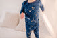 Child wearing long sleeved two piece pajama set on a dark blue background with constellations, galaxy, and stars in yellowish gold with blue cuffs 