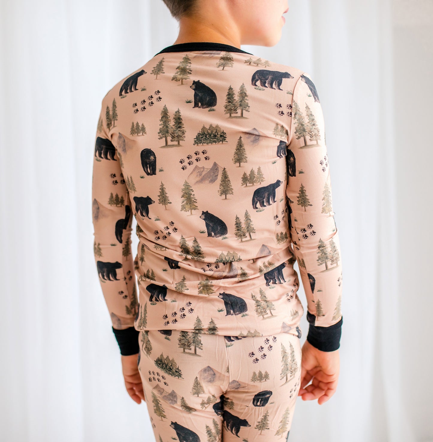Child wearing a long-sleeve shirt and pants with black bears, bear tracks, and forest pattern on a light brown background