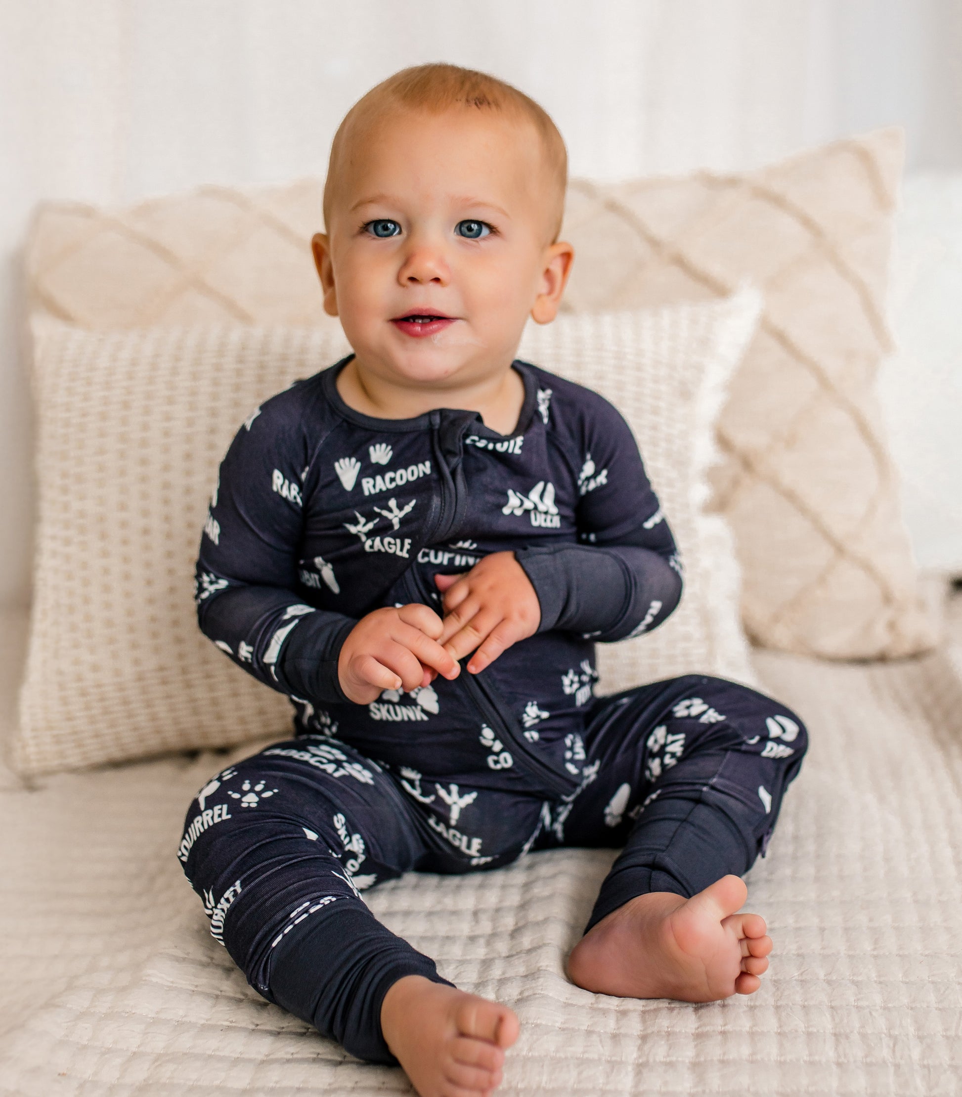 Baby wearing gray pajamas with white text and graphics, sitting on a beige couch