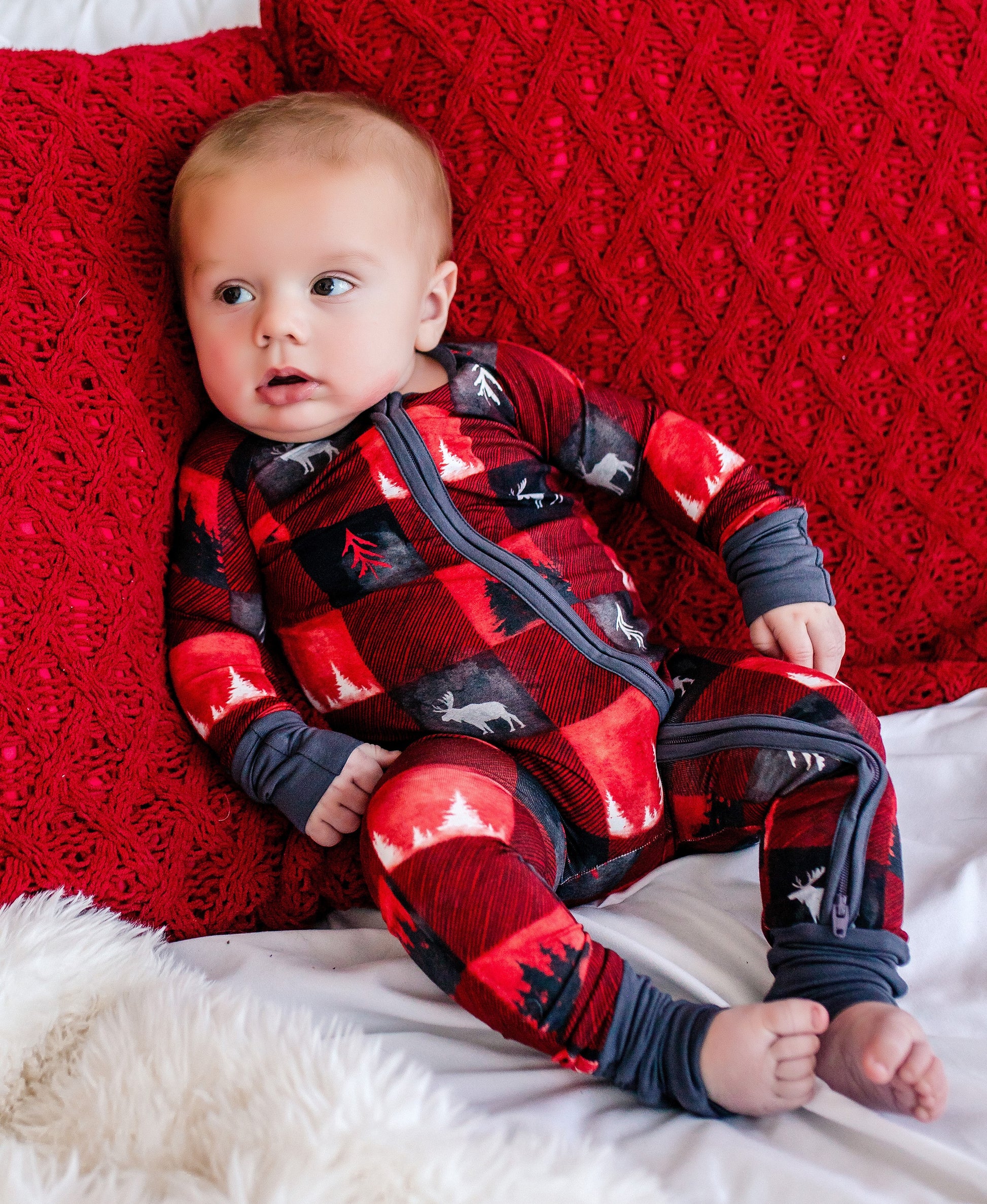Baby wearing a red and black plaid one piece with white moose and trees like the pine, fir, spruce, and maple, all native to the Maine woods. 