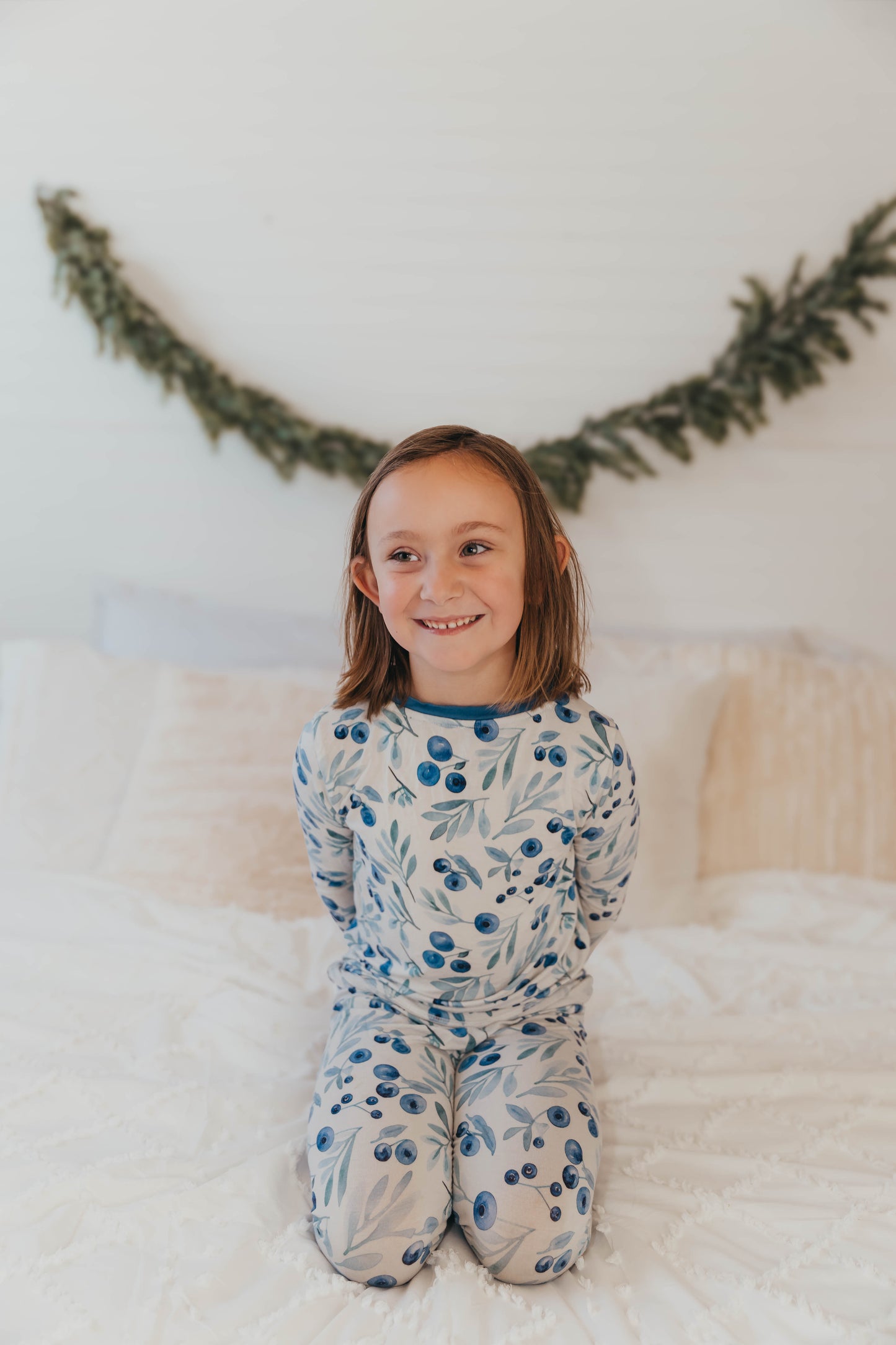 A child sitting on a bed wearing a two-piece pajama set with a floral pattern, predominantly blue and white. Maine wild blueberry two-piece pajama set for kids, Jaycob style, featuring a blueberry print inspired by Maine
