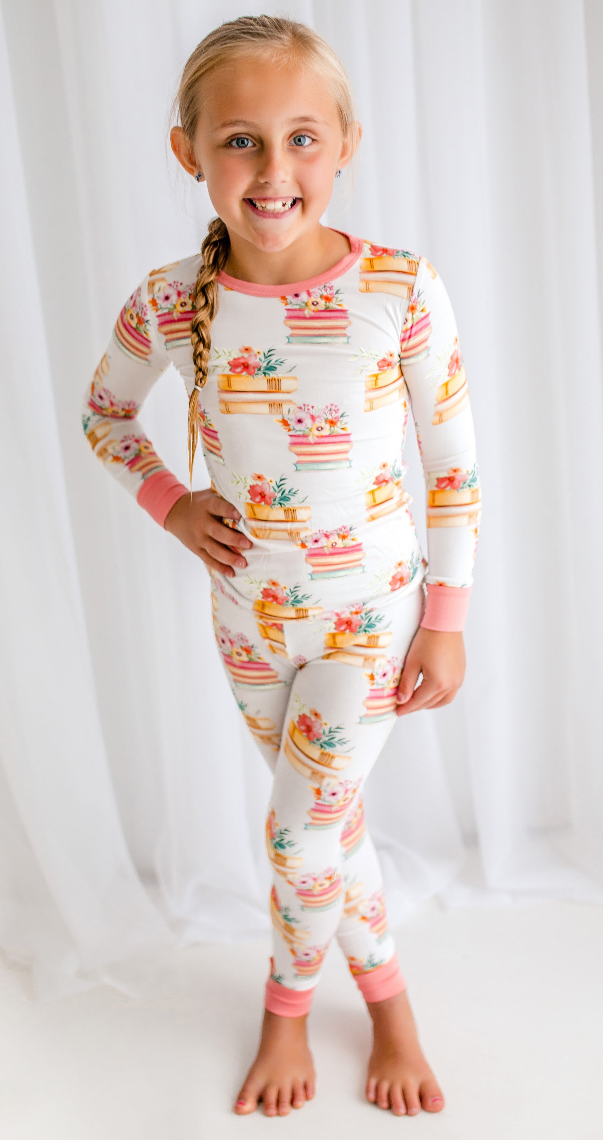 Young girl wearing a pajama set with book designs with flowers on the top of the stacked books on a white background