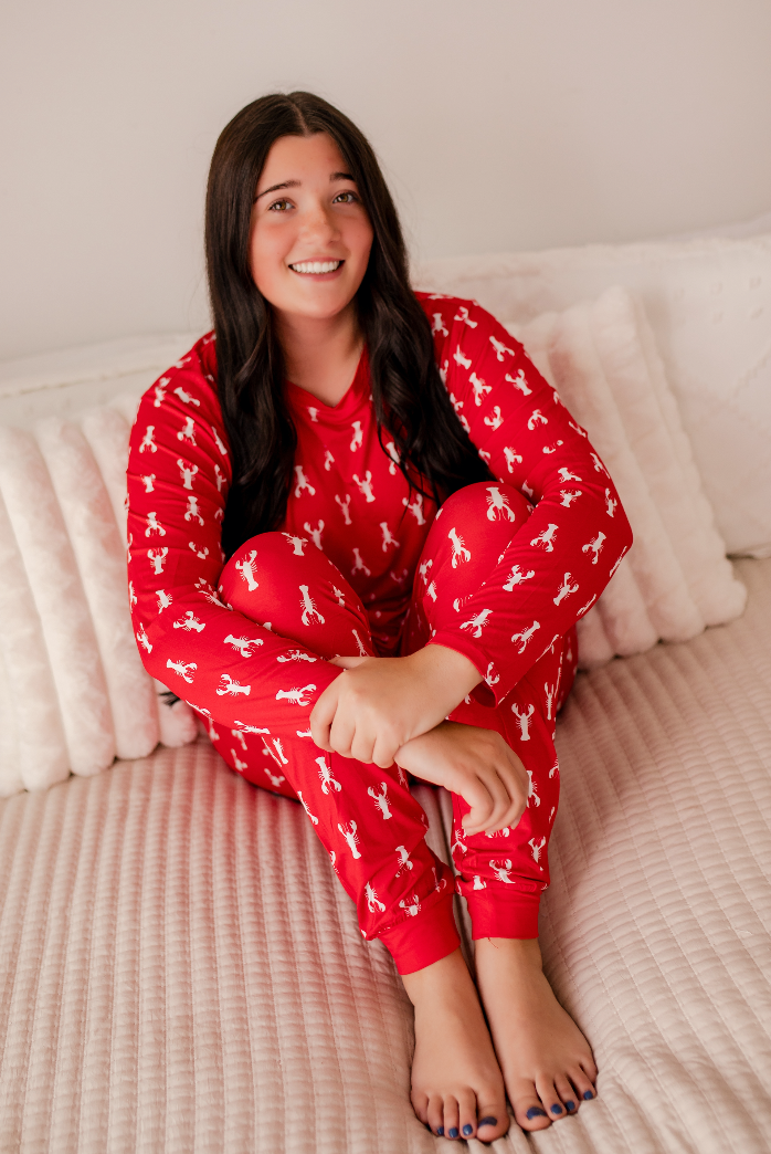 A smiling woman sits cross-legged on a bed wearing a red long-sleeve pajama set with a white allover bunny print. The room has a soft neutral background with a pinkish-beige textured bedspread and white pillows.