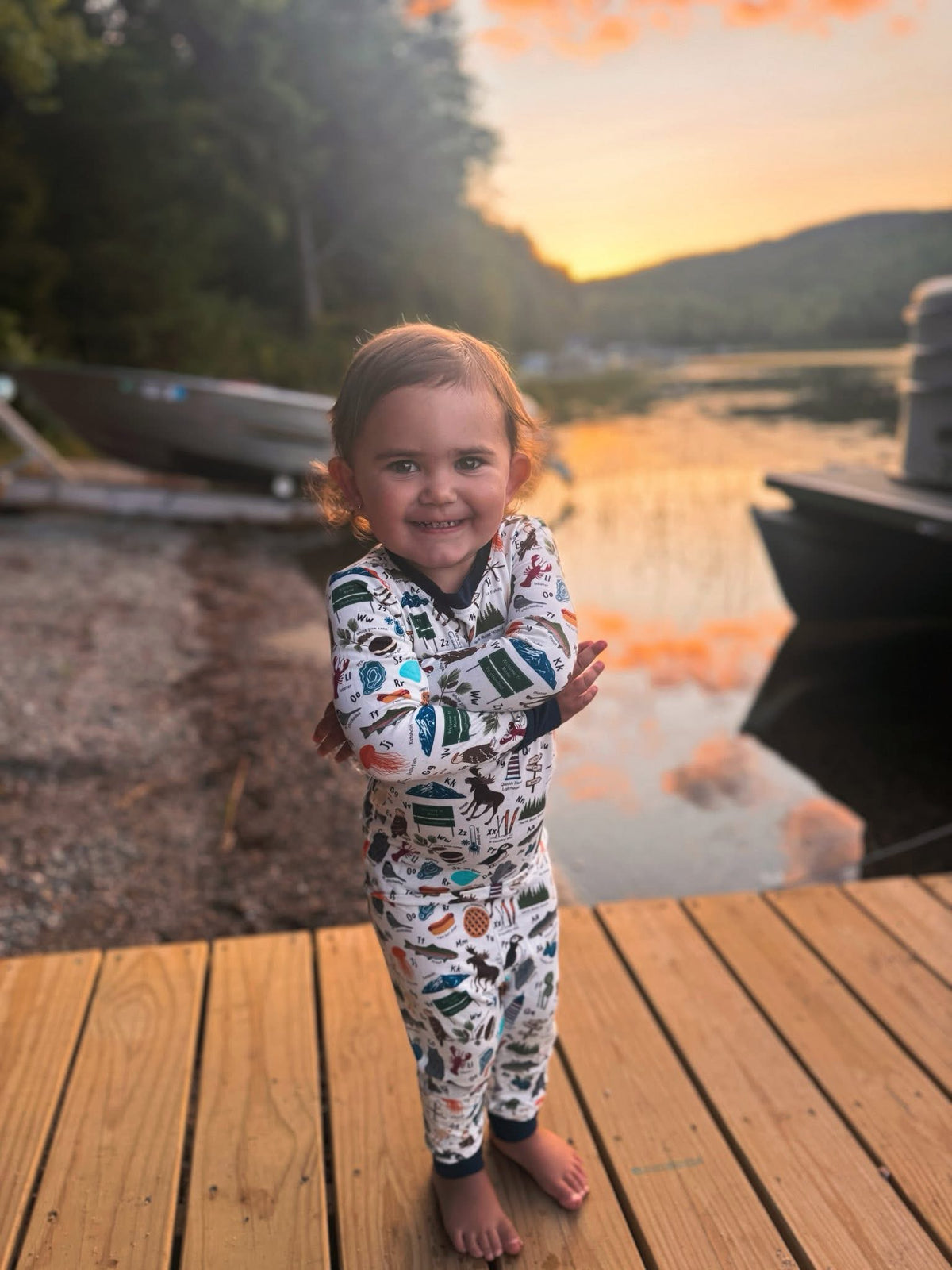 Child wearing an alphabet themed one piece pajama about all the things that represent maine, like puffins, moose, blueberries, eagles, and lobsters on a white background with blue cuffs. 
